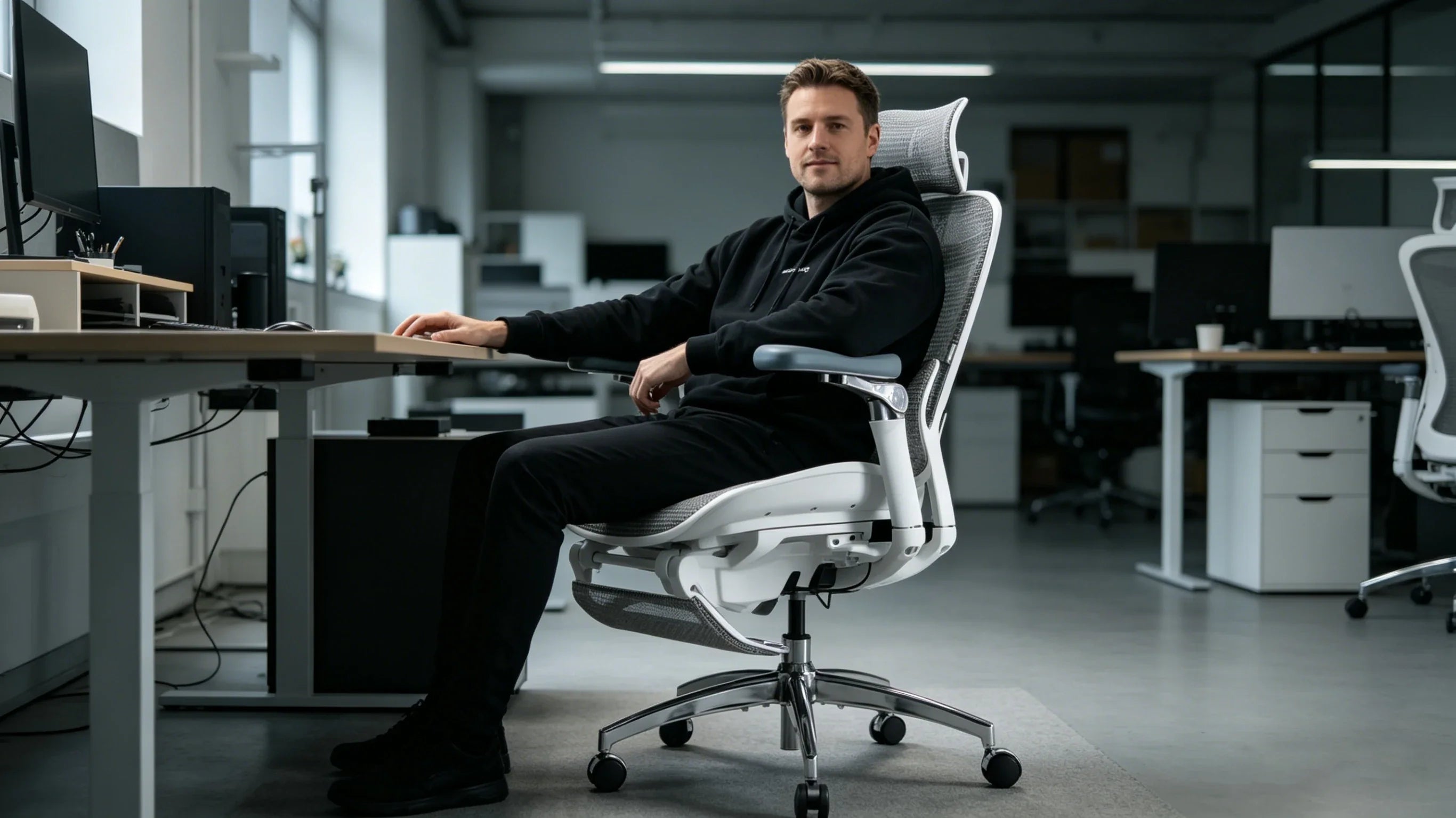 Full view of a 33-year-old male designer in a black sweatshirt sitting in the [desk chair adjustable height] in his tech-heavy studio.